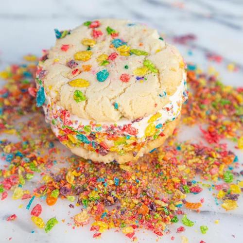 A colorful ice cream sandwich made with cookie layers and sprinkled with vibrant cereal bits on a marble surface.
