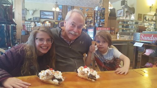 A smiling grandfather sits with two children, each holding ice cream sundaes, in a cozy shop filled with decor.