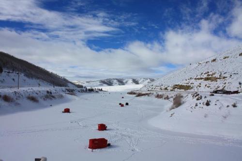 A snowy landscape with red ice fishing huts scattered across a frozen lake, surrounded by mountains and blue skies.