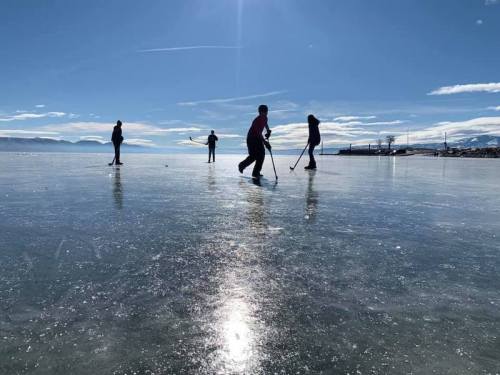 People playing hockey on a frozen lake under a clear blue sky with mountains in the background.