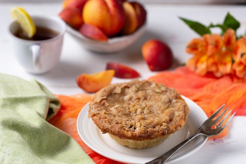 A slice of pie on a plate with a fork, surrounded by fresh peaches, a cup of tea, and colorful fabric.