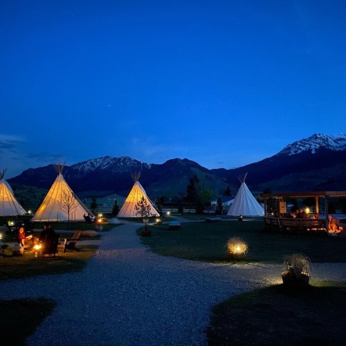 A serene night scene featuring illuminated tipis surrounded by mountains and a clear blue sky.