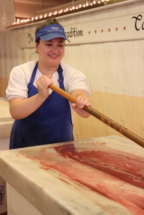 A smiling worker stirs a large batch of candy on a marble surface, wearing a blue apron and visor.