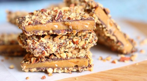 A stack of chocolate-covered toffee bars coated in crushed nuts, resting on a wooden surface.
