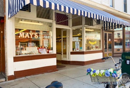 A storefront with "MAY'S" sign, featuring large windows and a blue and white striped awning, with a bicycle adorned with flowers.