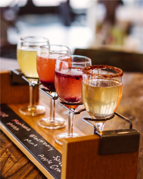 A wooden flight board holds four colorful cocktails in small glasses, showcasing various flavors and garnishes.