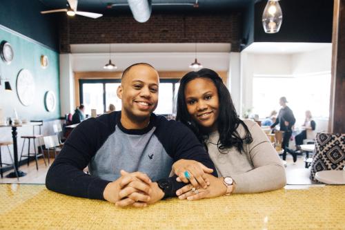 A smiling couple sits together at a café table, hands resting on the table, with a cozy atmosphere in the background.