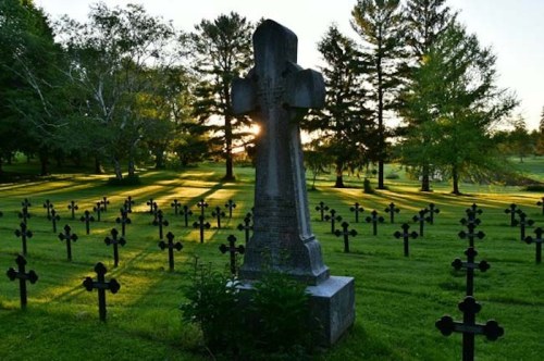 A serene cemetery scene with a large cross and rows of smaller crosses, illuminated by the setting sun through trees.