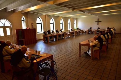 Monks seated at long tables in a spacious room, engaged in a meal, with large windows and a cross on the wall.