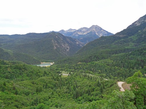 A panoramic view of lush green mountains and valleys under a cloudy sky, with a small lake visible in the distance.