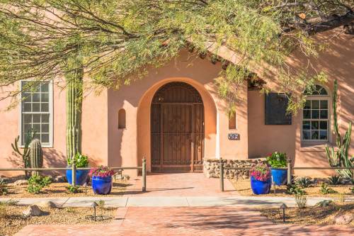 A southwestern-style home with a wooden door, surrounded by cacti and colorful blue planters filled with flowers.
