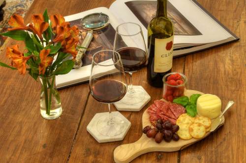 A wooden platter with cheese, salami, grapes, and crackers, two glasses of red wine, a bottle, and a vase of flowers.