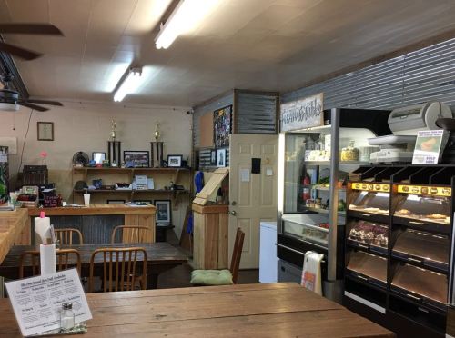 Interior of a cozy diner with wooden tables, a counter, trophies, and a display case filled with food.