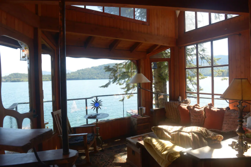 Cozy wooden cabin interior with large windows overlooking a serene lake and mountains, featuring comfortable seating.