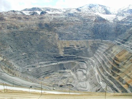 A large open-pit mine with layered rock formations and machinery, surrounded by mountains and a cloudy sky.