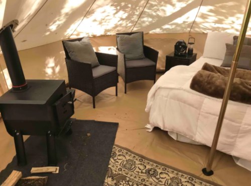 Cozy tent interior with a bed, two chairs, a small table, and a wood stove, surrounded by warm lighting.