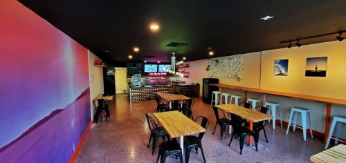 Interior of a modern restaurant with wooden tables, bar area, and colorful wall art. Bright and inviting atmosphere.