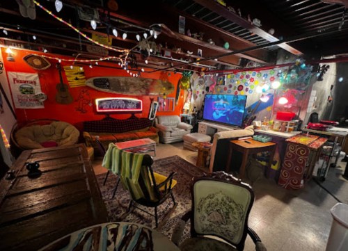 A colorful, eclectic lounge with cozy seating, vibrant wall art, and a TV, illuminated by string lights.