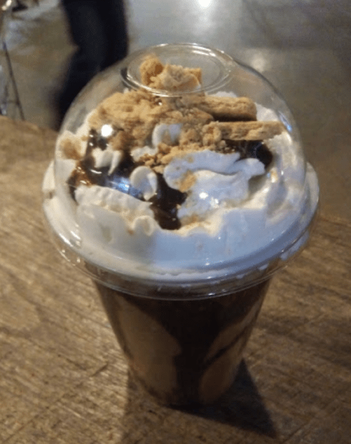 A dessert drink in a clear cup topped with whipped cream, chocolate drizzle, and crushed cookies.