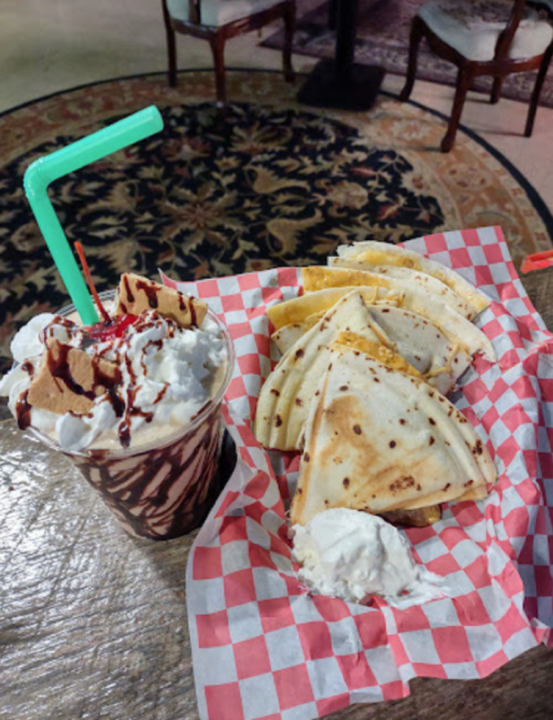 A dessert plate featuring a chocolate milkshake with whipped cream and a plate of quesadillas on a checkered paper.