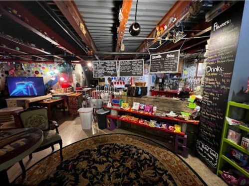 A colorful basement shop with a menu board, snacks, drinks, and a cozy seating area, featuring vibrant decor and lighting.