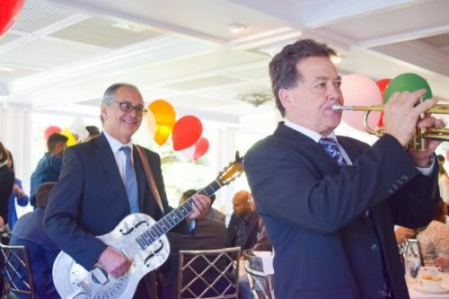 Two musicians perform at a lively event, one playing a guitar and the other a trumpet, with colorful balloons in the background.