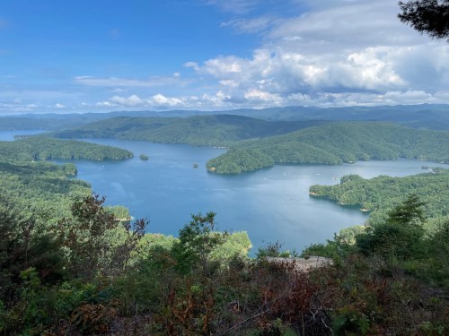 A scenic view of a lake surrounded by lush green hills under a partly cloudy sky.