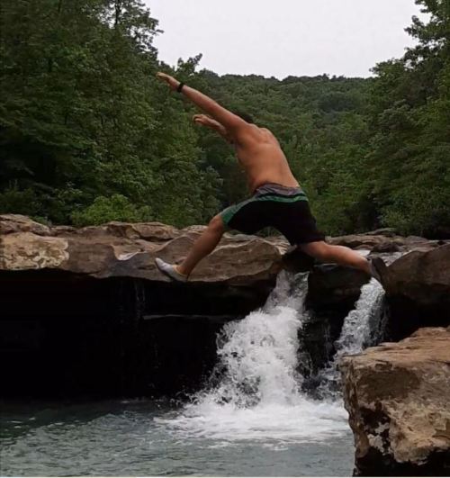 A person jumps off a rock into a waterfall surrounded by lush greenery.