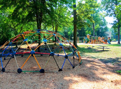 A colorful climbing dome in a playground surrounded by trees, with a play structure and picnic area in the background.