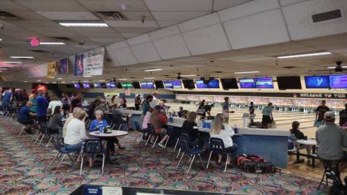 A busy bowling alley with people bowling, sitting at tables, and colorful decor on the walls.