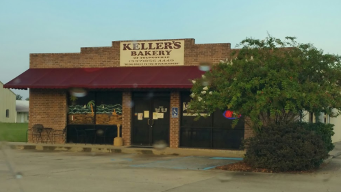 A brick bakery building with a red awning, sign reading "Keller's Bakery," and a small tree in front.