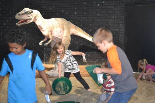 Children playing in a dinosaur-themed exhibit, digging in sand with a dinosaur model in the background.