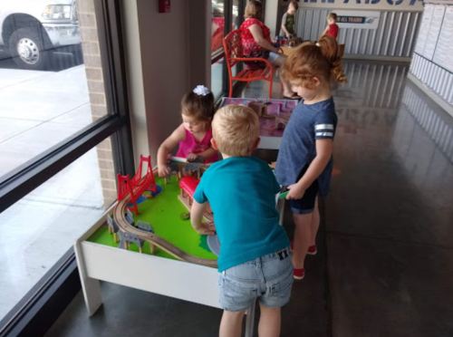 Three children play with a colorful train set at a table near a window, with adults seated in the background.