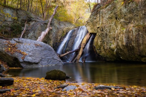 A serene waterfall cascades over rocks, surrounded by autumn foliage and a calm pool reflecting the landscape.