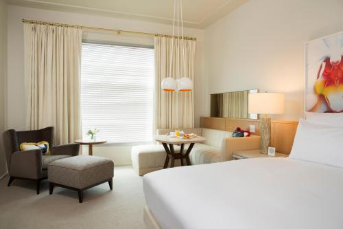 A modern hotel room featuring a bed, seating area, table, and large window with curtains. Bright and inviting decor.