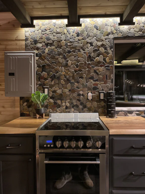 A modern kitchen featuring a stone backsplash, stainless steel stove, wooden countertop, and subtle lighting.