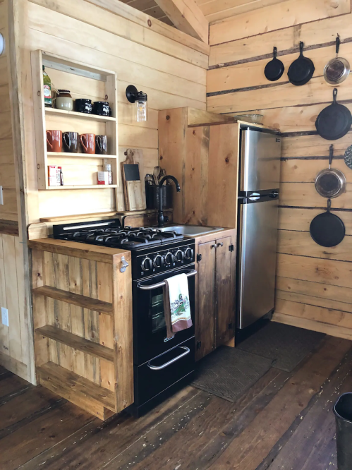 Cozy kitchen with wooden walls, a black stove, stainless steel fridge, and open shelving with mugs and cookware.