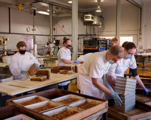 Workers in a bakery wearing masks and gloves, preparing and packaging baked goods in a clean, bright kitchen.