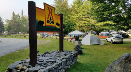 A KOA campground sign with tents and campers in the background, surrounded by trees and rocky landscaping.