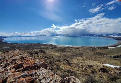 A panoramic view of a serene blue lake surrounded by rocky terrain and distant mountains under a bright sky.