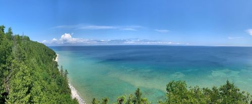 A panoramic view of a clear blue lake bordered by lush green trees under a bright blue sky.