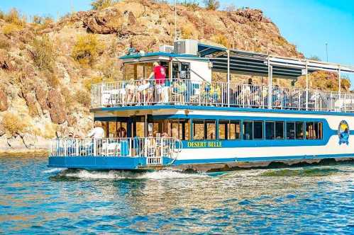 A blue and white tour boat, the Desert Belle, cruises on a lake surrounded by rocky hills and clear blue skies.