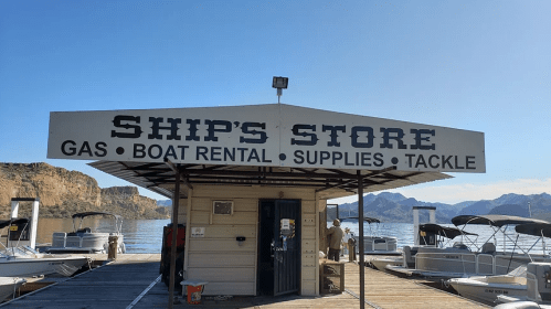 A boat rental and supply store on a dock, with a sign reading "Ship's Store" and scenic mountains in the background.