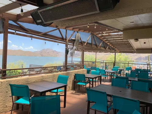 A lakeside restaurant patio with turquoise chairs and tables, featuring a scenic view of mountains and water.