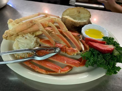 A plate of crab legs, a baked potato, strawberries, kale, and a small dish of olive oil on a restaurant table.