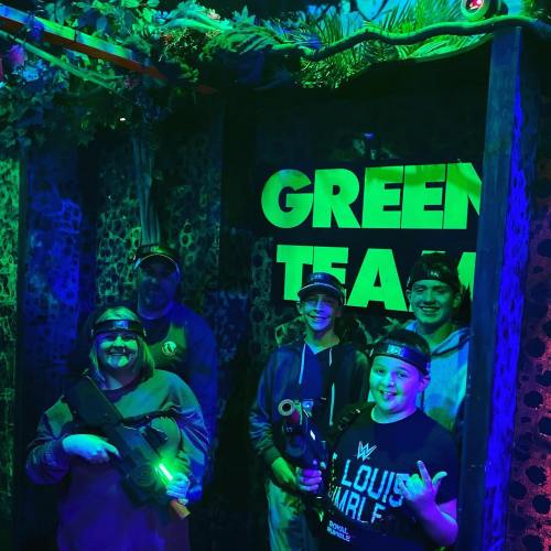 A group of four people in a dark, neon-lit room, wearing headsets and holding laser tag equipment, smiling for the camera.