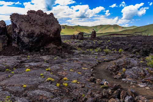 Lava rock formations with a winding path, set against a backdrop of green hills and a blue sky with clouds.
