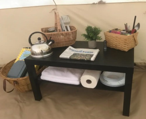 A black table in a tent, holding a kettle, baskets of utensils, a newspaper, and towels.