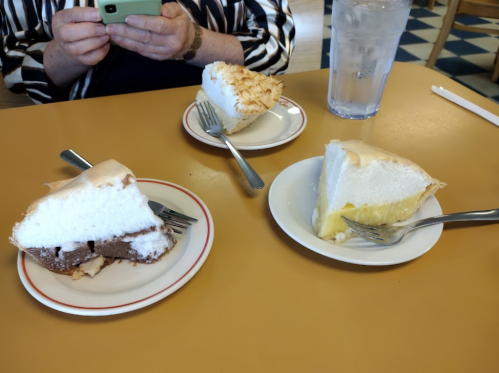 Three slices of pie on a table: chocolate, lemon, and coconut meringue, with a glass of water in the background.