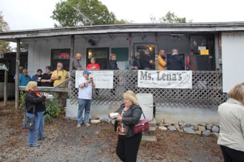 A crowd gathers outside Ms. Lena's restaurant, with people chatting and holding food containers on a cloudy day.